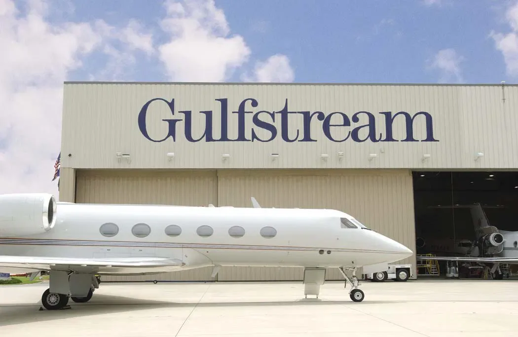 A white Gulfstream business jet parked on the apron outside a hangar with "Gulfstream" lettered in large blue type across the front.