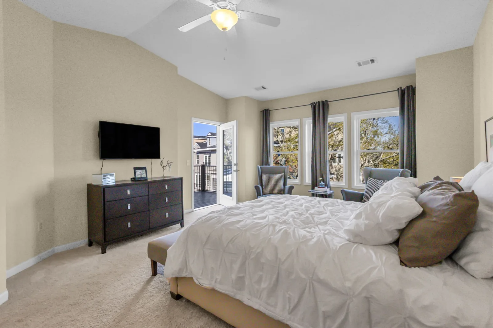 Carpeted primary bedroom with a vaulted ceiling and ceiling fan, king bed in white bedding, wall-mounted TV above a dark dresser, and French doors opening to a small balcony beside three curtained windows.