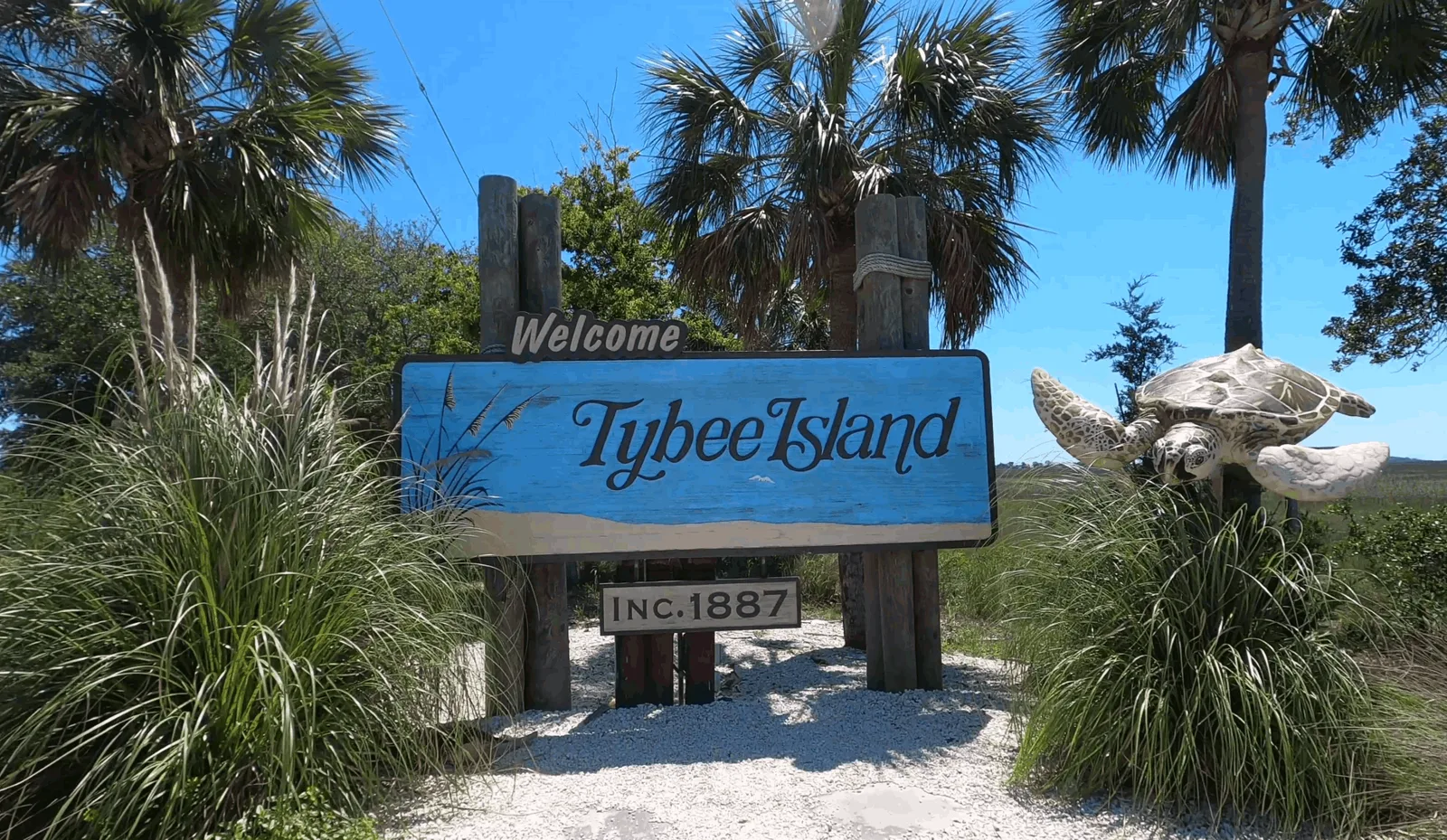 Weathered blue "Welcome Tybee Island" sign marked Inc. 1887, framed by palmettos and a carved sea turtle.